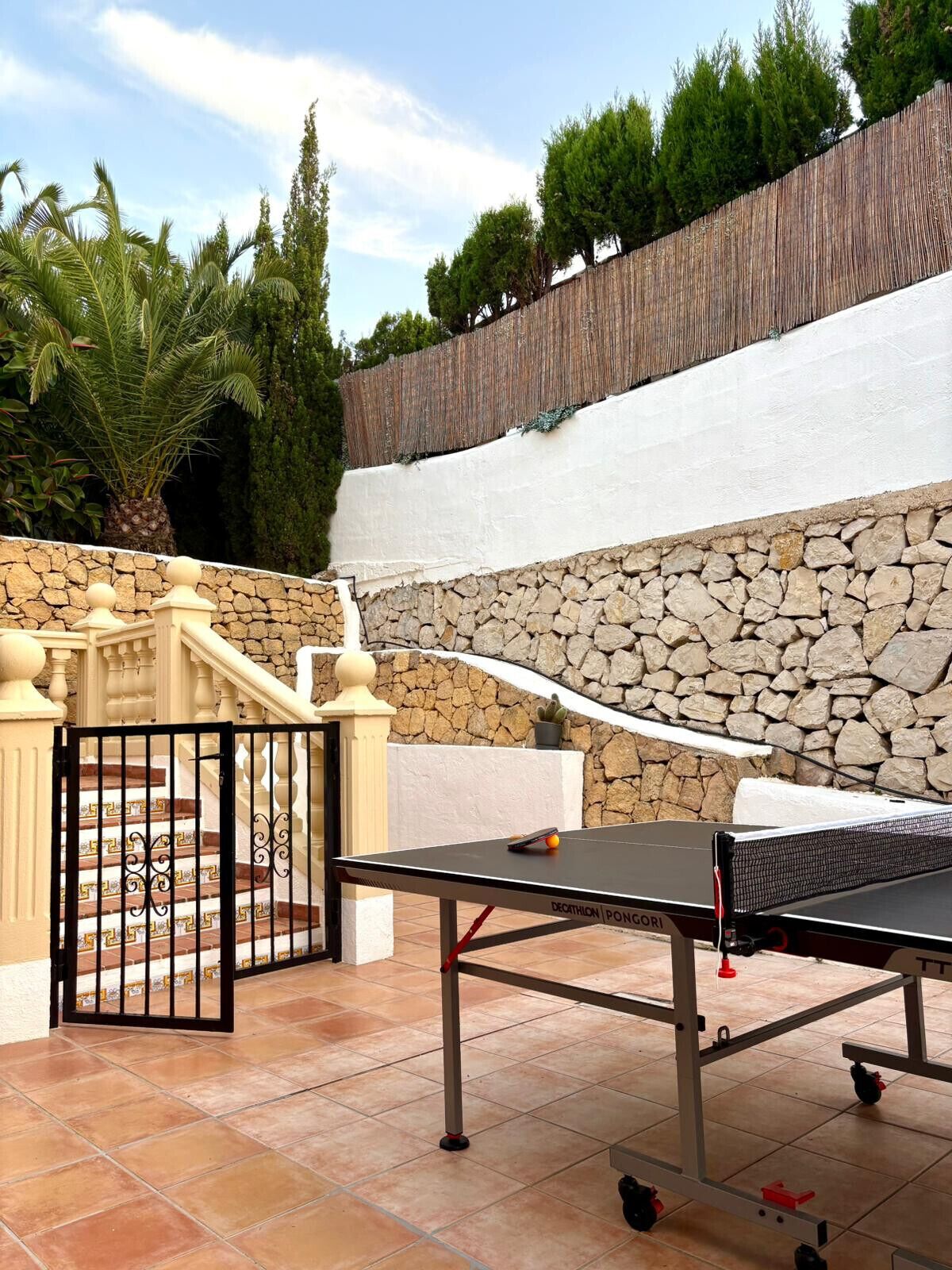 An outdoor ping pong table set up on a patio with a stone wall and a wooden fence in the background.