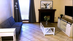 Una sala de estar con un sofá, una mesa de centro blanca, una chimenea con un cuadro enmarcado y un televisor sobre un mueble.