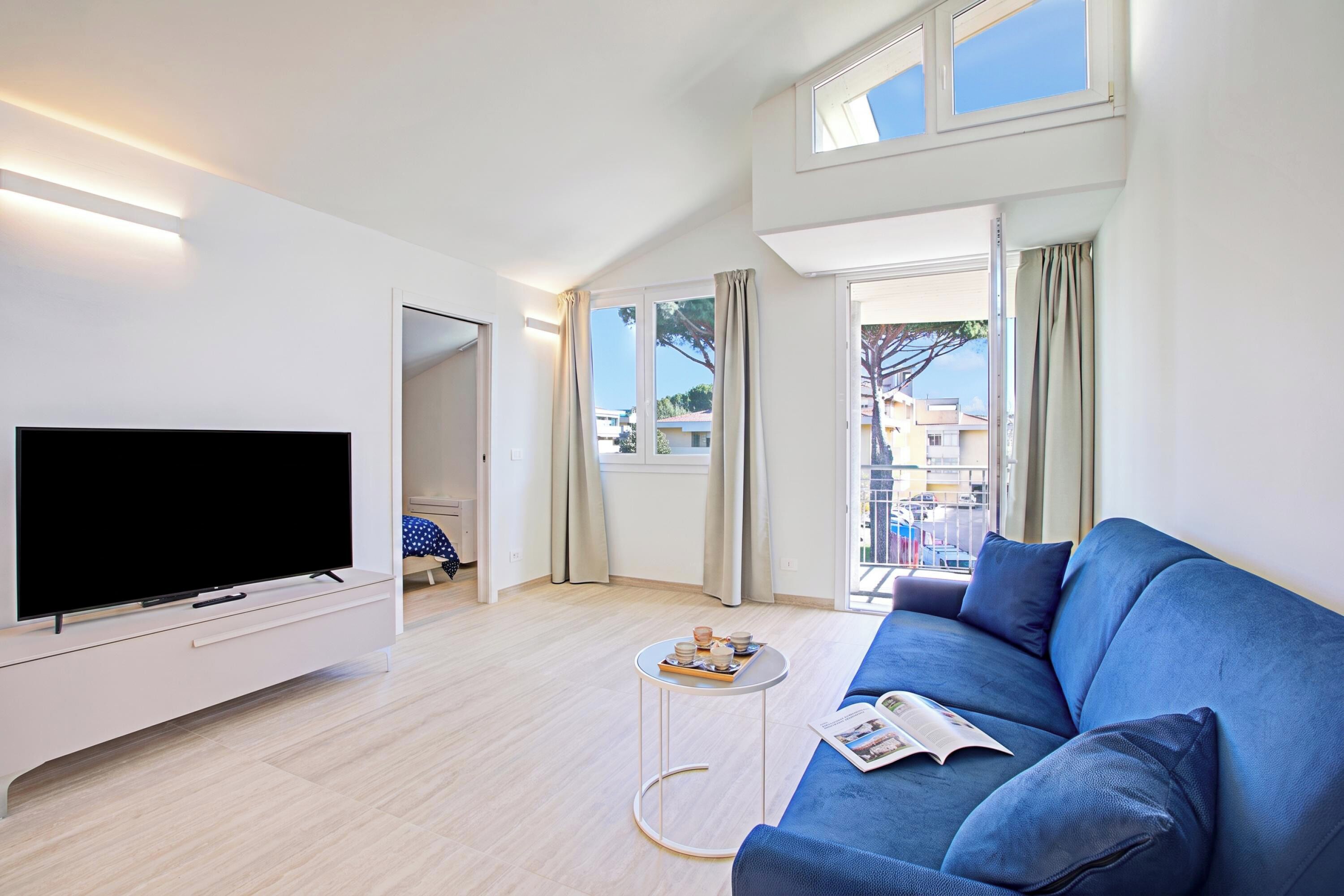 A modern living room with a flat-screen TV, a blue sofa, a coffee table, and a balcony with curtains.