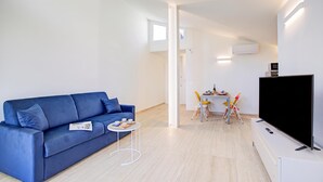 Un salon moderne avec un canapé bleu, une petite table ronde sur laquelle se trouve un plateau avec des tasses, une table à manger avec des chaises et un téléviseur à écran plat posé sur un meuble.