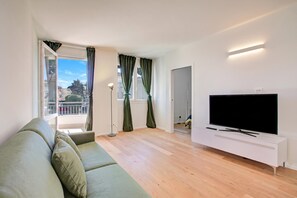A modern living room with a green sofa, a flat-screen TV on a white stand, and a balcony with curtains.