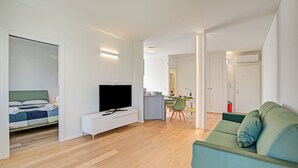 Una sala moderna con una televisión de pantalla plana, un sofá verde, un comedor y una cocina al fondo, sobre piso de madera.