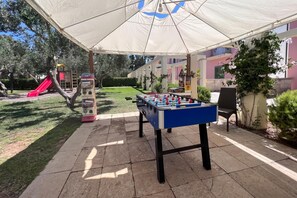 Un patio exterior con mesa de fútbol, zona de juegos con tobogán y un área de estar techada.