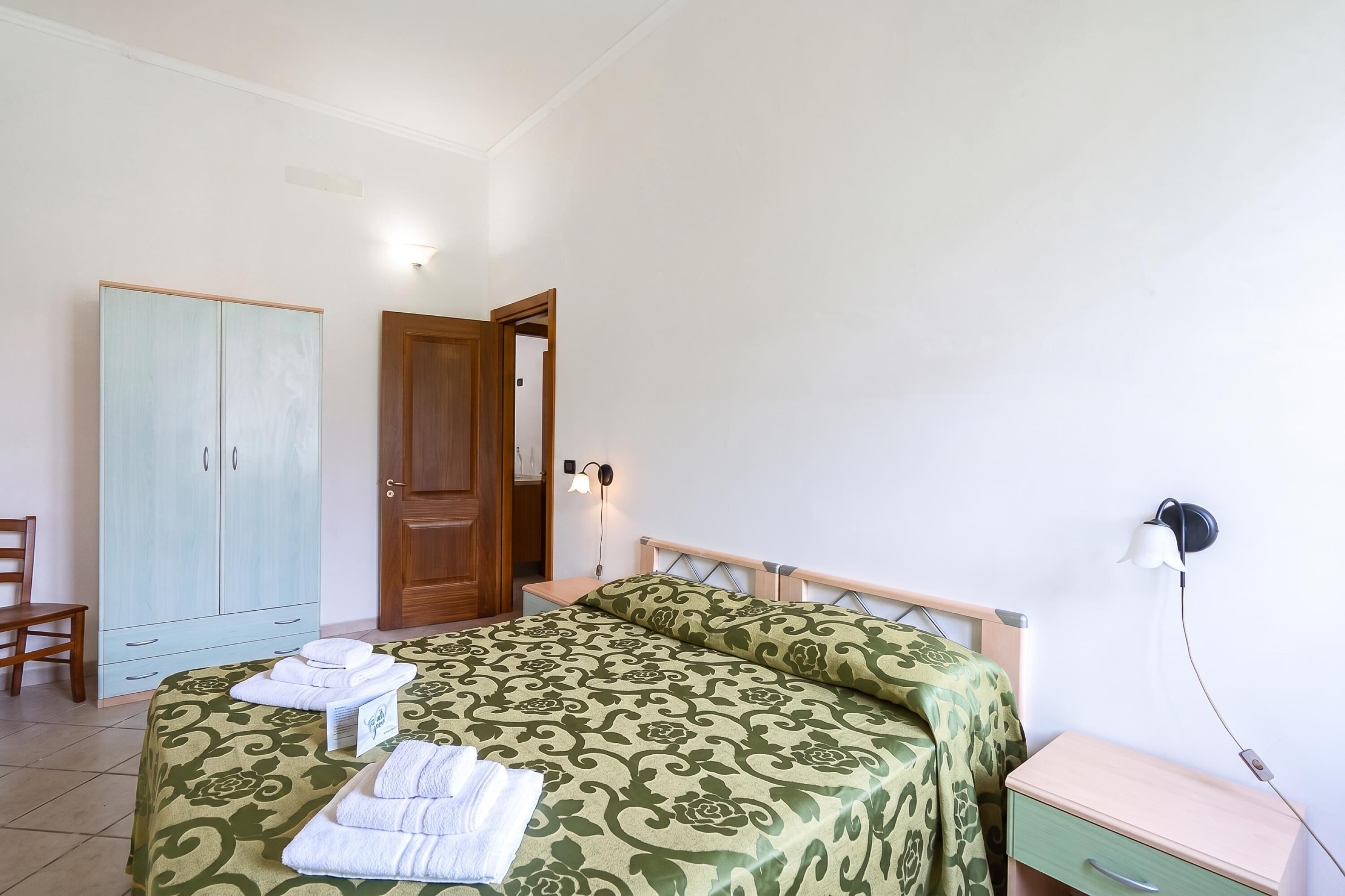 Un dormitorio con cama, armario de madera, mesita de noche, una silla y una lámpara de pared.