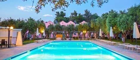 A swimming pool with lounge chairs and umbrellas, surrounded by trees and a building with pink walls.