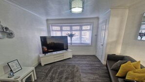 Un salon moderne avec un téléviseur à écran plat posé sur un meuble TV blanc, un canapé marron foncé agrémenté de coussins jaunes, un tapis à motifs et un luminaire au plafond.