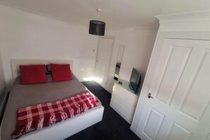 Un dormitorio con una cama, dos almohadas rojas, una colcha roja y blanca, un televisor sobre una cómoda y una puerta blanca.