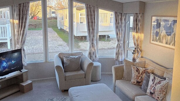Un salon moderne comprenant un canapé, un fauteuil et un pouf, un téléviseur à écran plat et de grandes fenêtres avec des rideaux.