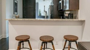 A modern kitchen with a central island, three wooden stools, pendant lighting, and a view into the dining area. - Urban King Loft - Cali King - 8 Min to Broadway! (Nashville)