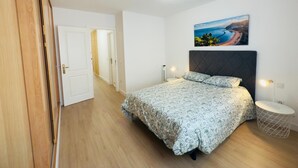 A bedroom with a bed, a nightstand, a wall-mounted lamp, and a painting of a beach scene.