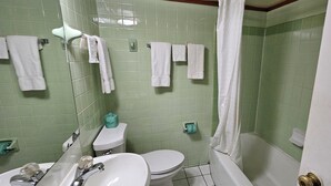 A bathroom with green tiled walls, a white toilet, a sink, and a bathtub. There are several white towels hanging on a rack. - Deluxe Inn (Stuttgart)