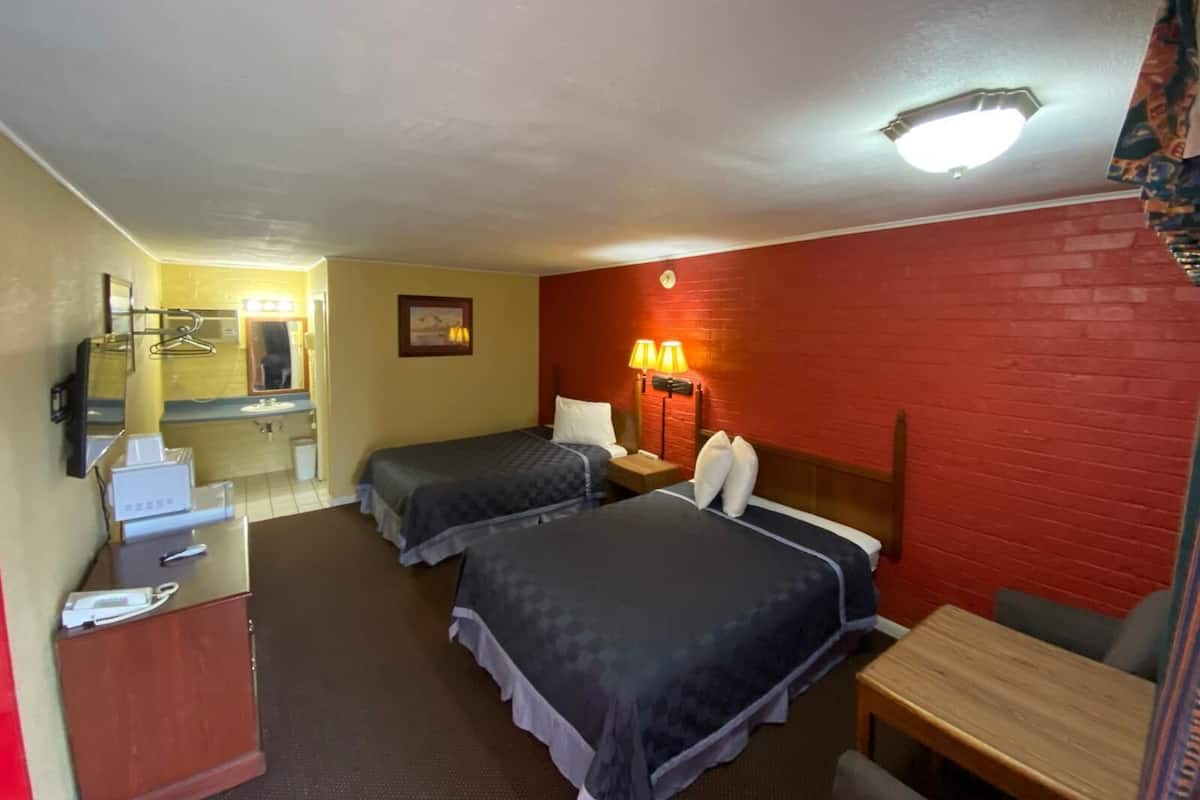 Ett hotellrum med två sängar, ett skrivbord, en stol, ett litet bord, en spegel, en lampa och en vägg av rött tegel.