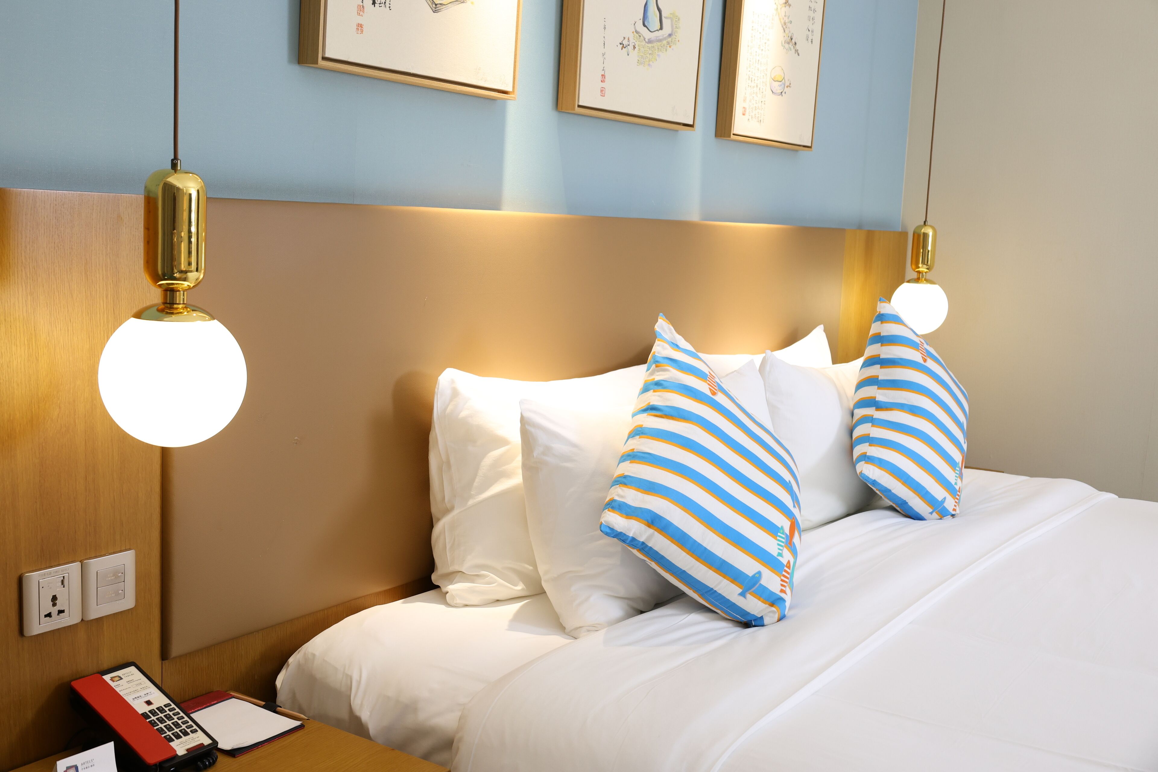 A hotel room with a neatly made bed, a bedside table with a telephone and a red book, and wall-mounted artwork above the bed.