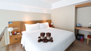 Une chambre d’hôtel avec un grand lit, un bureau, une chaise, une table de chevet avec une lampe, un téléphone et un ours en peluche.