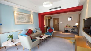 Une chambre d’hôtel moderne dotée d’un espace salon comprenant un canapé, des fauteuils, une table basse et un téléviseur. On y trouve également un bureau avec une chaise et un miroir en arrière-plan.
