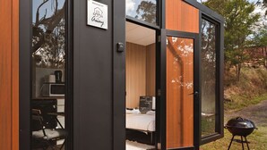 A modern, compact cabin with a wooden exterior and glass doors, featuring a bed, a small kitchenette, and a barbecue grill outside. - Maroota Greens JOY by Tiny Away (Maroota)