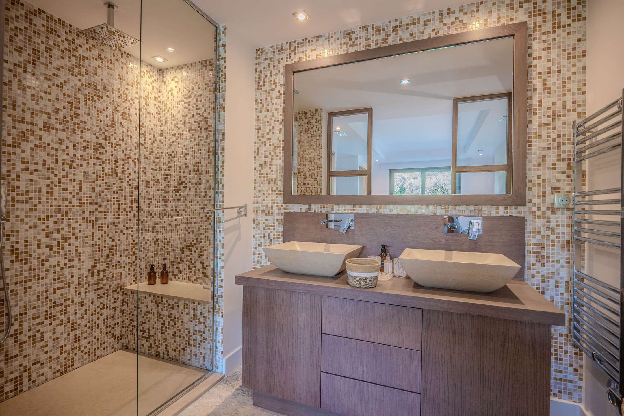 Een moderne badkamer met mozaïektegel wanden, twee wasbakken op consoles, een grote spiegel en een douche met glazen wanden.