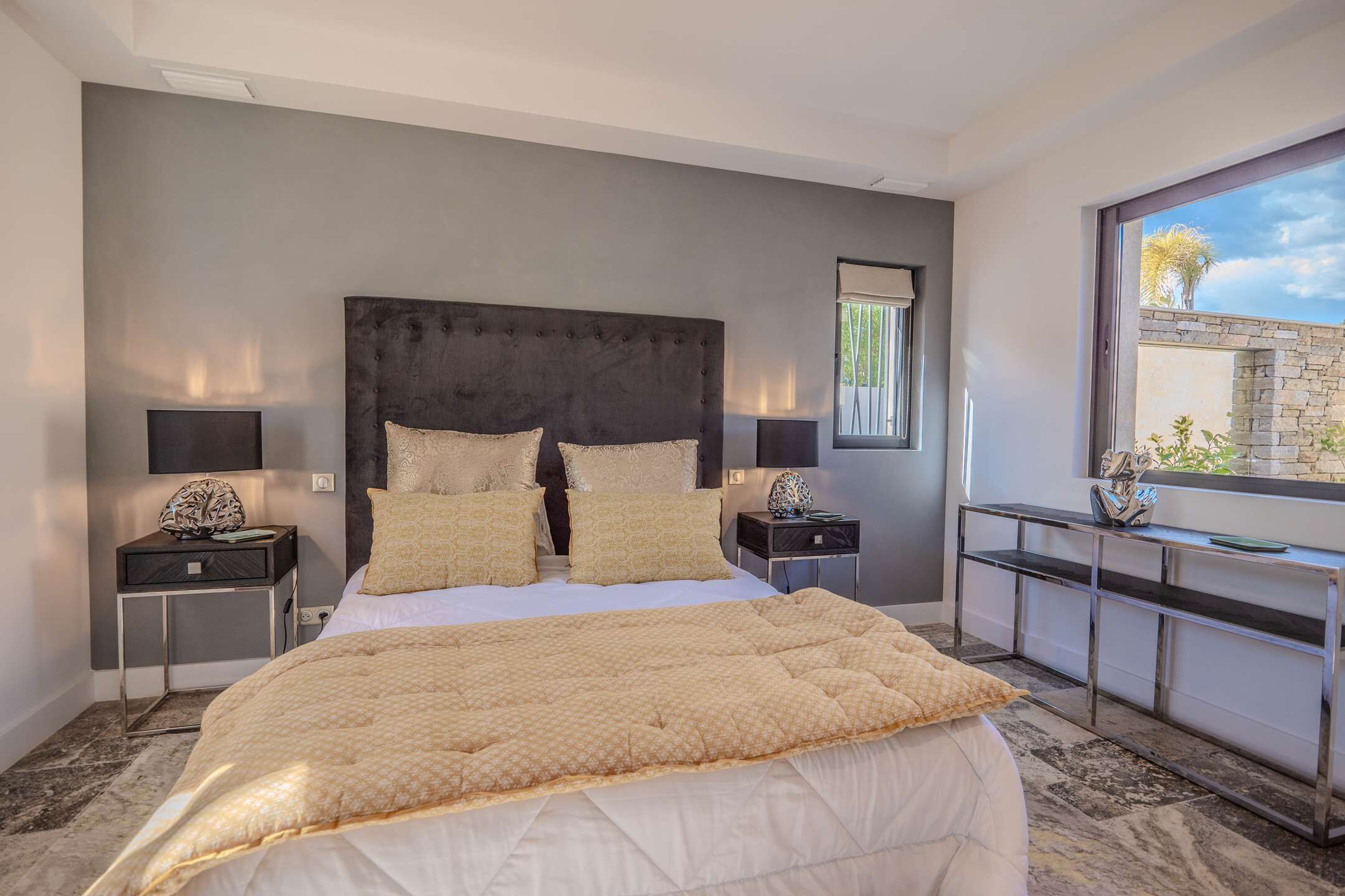 A modern hotel room with a large bed, two bedside tables with lamps, a console table, and a view of the outdoors.