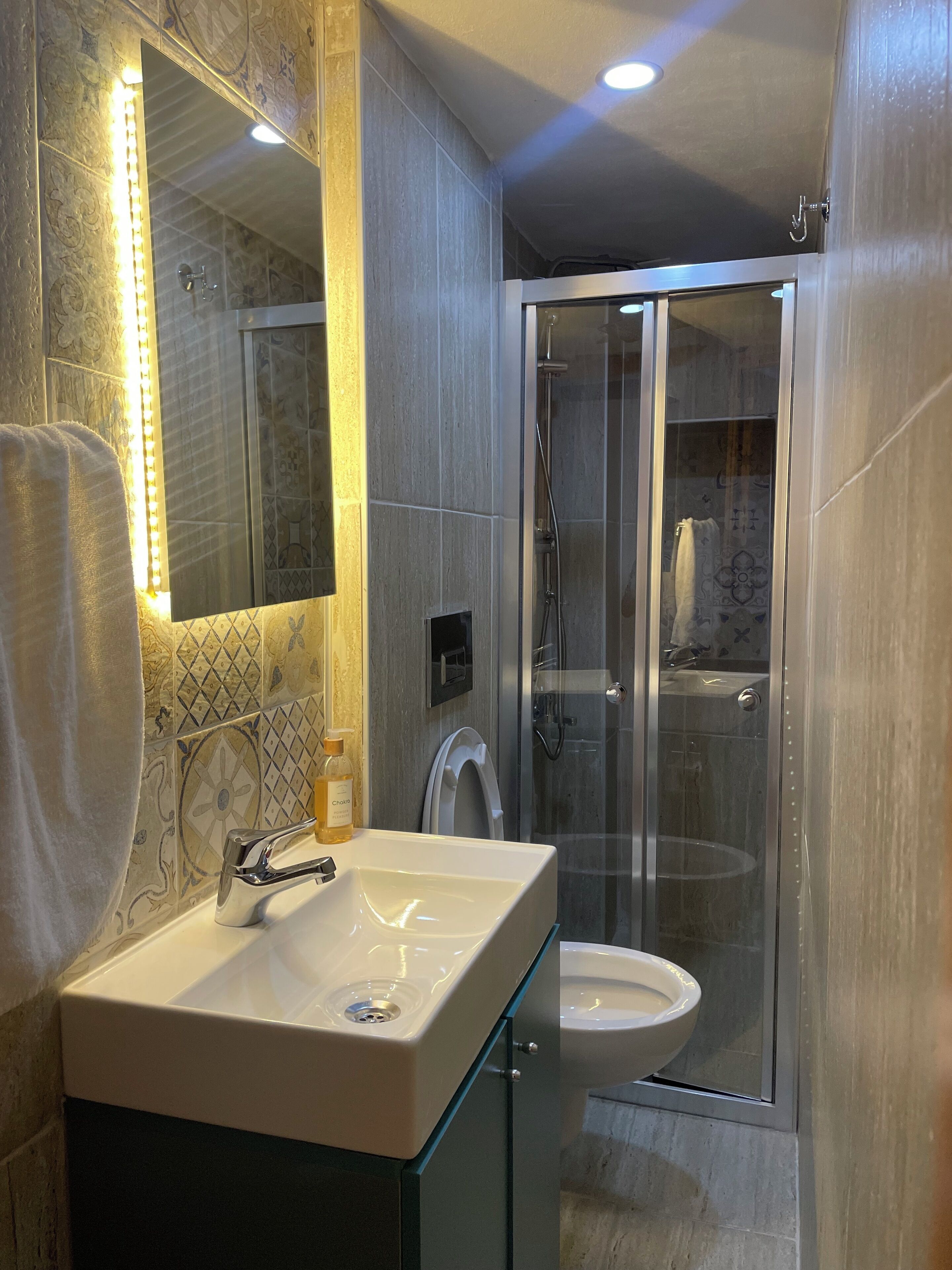 A modern bathroom with a glass shower enclosure, a white sink, and a mirror with LED lights.