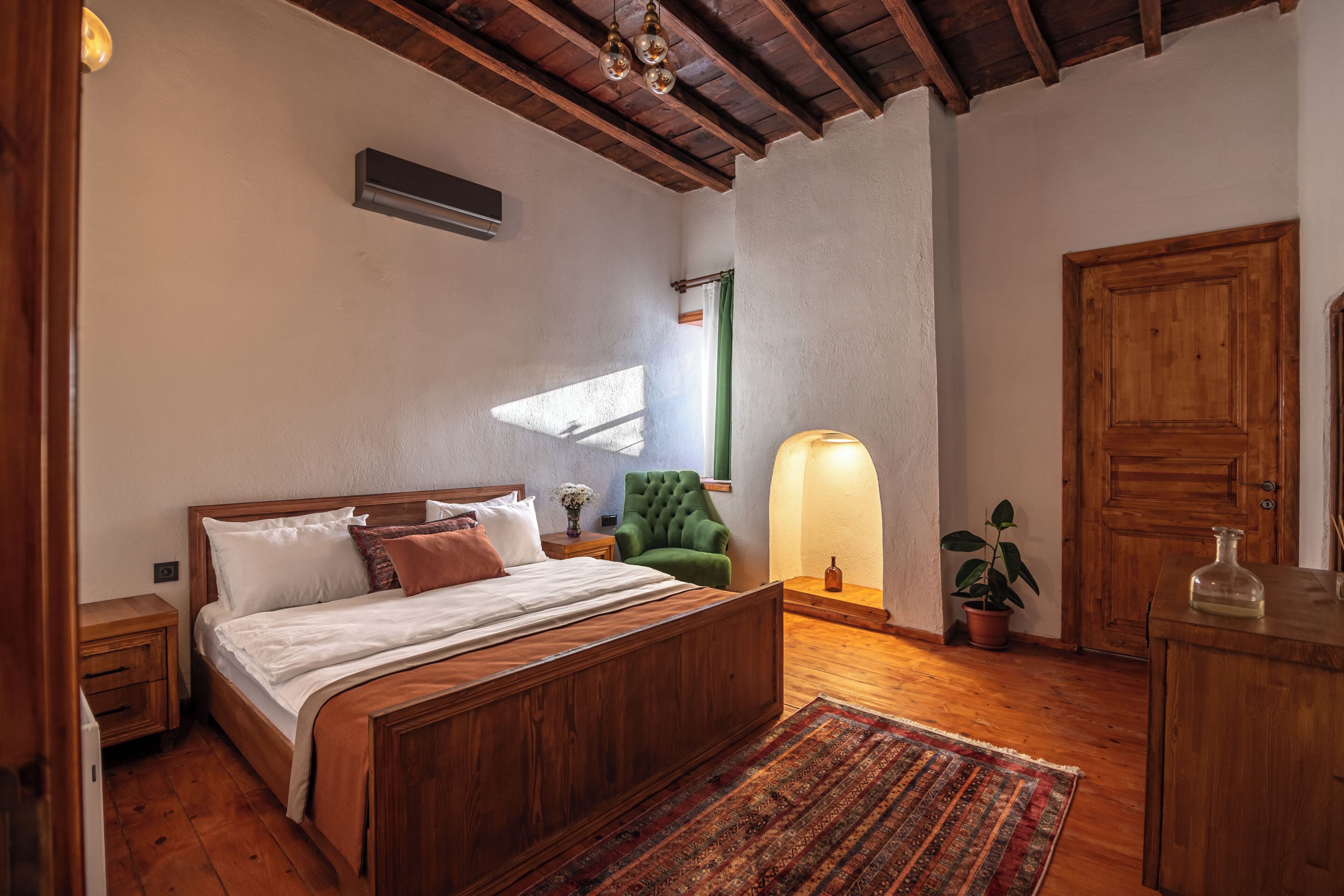 Quarto com cama de madeira, poltrona verde, mesa de cabeceira, abajur, planta em vaso e guarda-roupa de madeira.