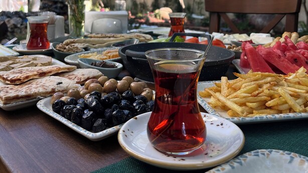 A table set with various dishes including flatbread, olives, and a glass of tea, with a view of trees and a building in the background. - Marassa Konak Otel (Ermenek)