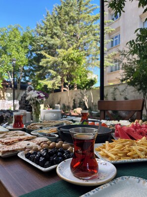 A table set with various dishes including flatbread, olives, and a glass of tea, with a view of trees and a building in the background. - Marassa Konak Otel (Ermenek)