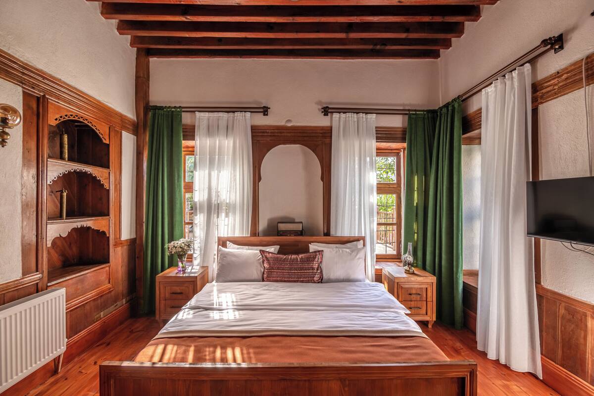 A wooden-framed bedroom with a bed, nightstands, a TV, and a window with curtains.