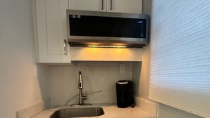 Cocina compacta con armarios blancos, un lavamanos de acero inoxidable, una cafetera negra y un microondas sobre la encimera.