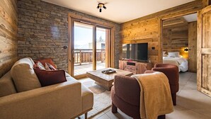 A cozy living room with a stone wall, wooden paneling, a sofa, armchairs, a coffee table, and a TV. - 6-person flat in a new residence on the snow front (LA TOUSSUIRE)