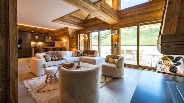 A cozy cabin interior with a living area featuring a sofa, ottoman, and armchairs, a kitchen, and large windows offering a view of the outdoors. - 12-person flat with jacuzzi and sauna on the snow front (LA TOUSSUIRE)