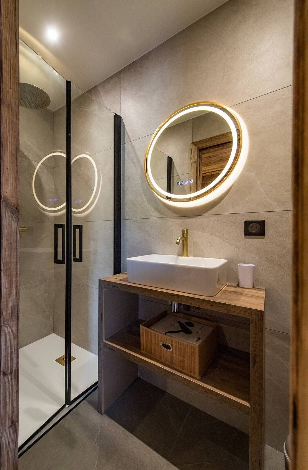 A modern bathroom with a glass shower enclosure, a white sink with a gold faucet, and a wooden vanity with a mirror.