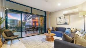 A modern living room with a sofa, armchair, and coffee table, a glass door leading to an outdoor area, and a painting on the wall. - Convenient CBD Location, Pool/Gym + walk to shops (Darwin)