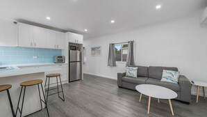 Eine moderne Küche mit weißen Schränken, einem Edelstahlkühlschrank und einer dunkelblauen Küchenwand. Im Wohnbereich befindet sich ein graues Sofa mit blauen Kissen und ein kleiner weißer Tisch.