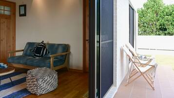 Una sala de estar acogedora con un sofá azul, un reposapiés estampado y una mesa de centro de madera. Un ventanal corredera da a un área exterior con una terraza de madera y una silla.