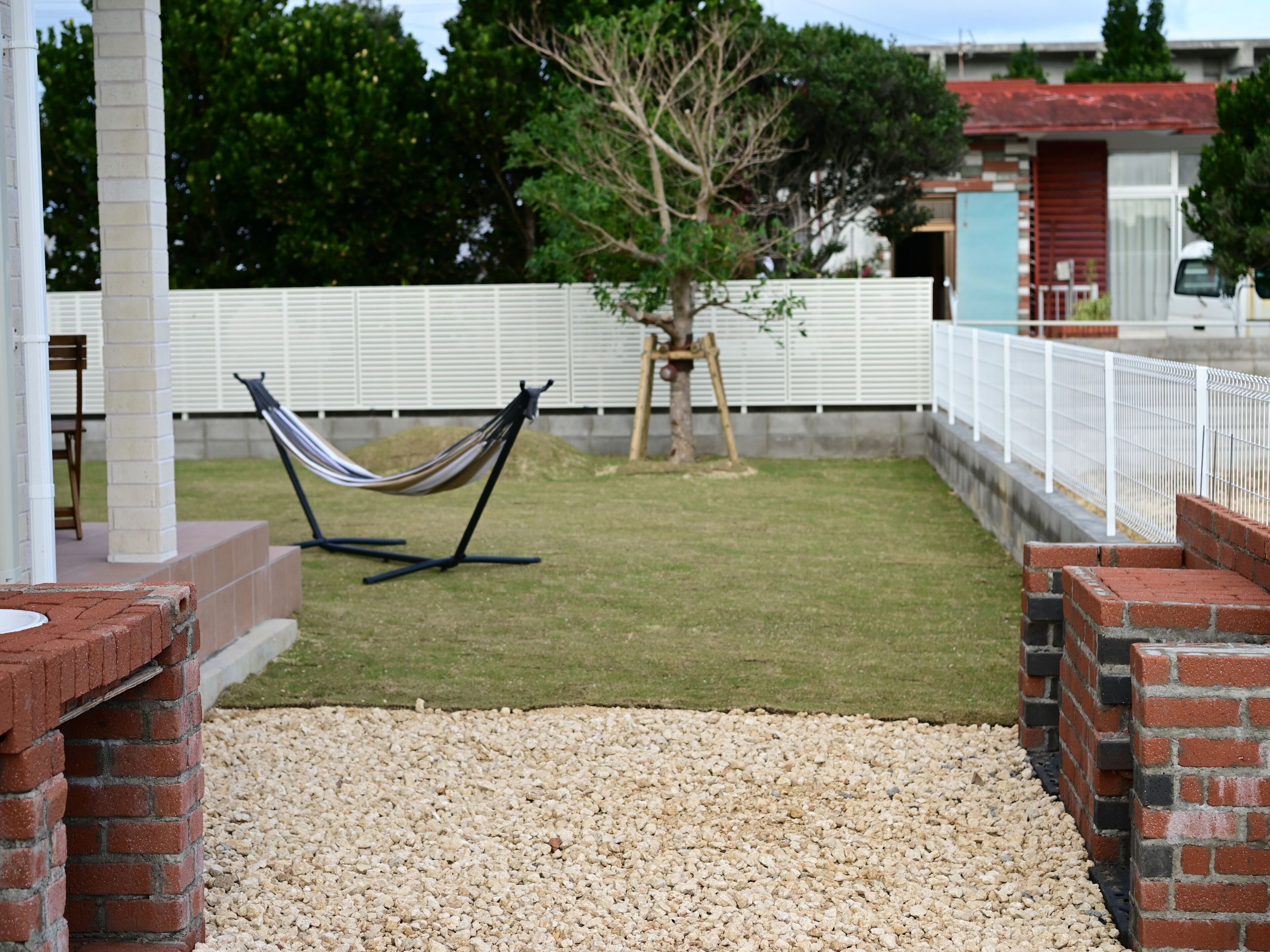 A hammock on a brick patio with a white fence and a tree in the background.