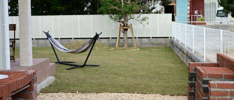 A hammock on a brick patio with a white fence and a tree in the background.