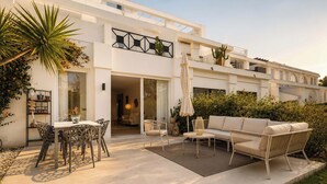 A modern house with a patio area featuring a dining table, chairs, a sofa, and a small table with a lamp. - Las Terrazas 2 | Modern Golf Terrace Pool (Benahavís)