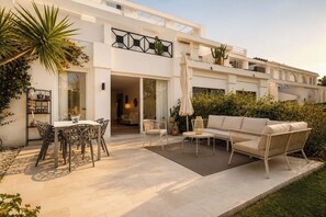 A modern house with a patio area featuring a dining table, chairs, a sofa, and a small table with a lamp.