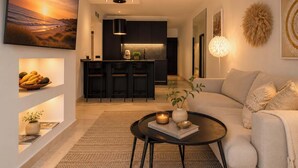 A modern living room with a sofa, a coffee table, and a kitchen area in the background. - Las Terrazas 2 | Modern Golf Terrace Pool (Benahavís)