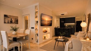 A modern living room with a dining area, a kitchen, and a TV mounted on the wall. - Las Terrazas 2 | Modern Golf Terrace Pool (Benahavís)