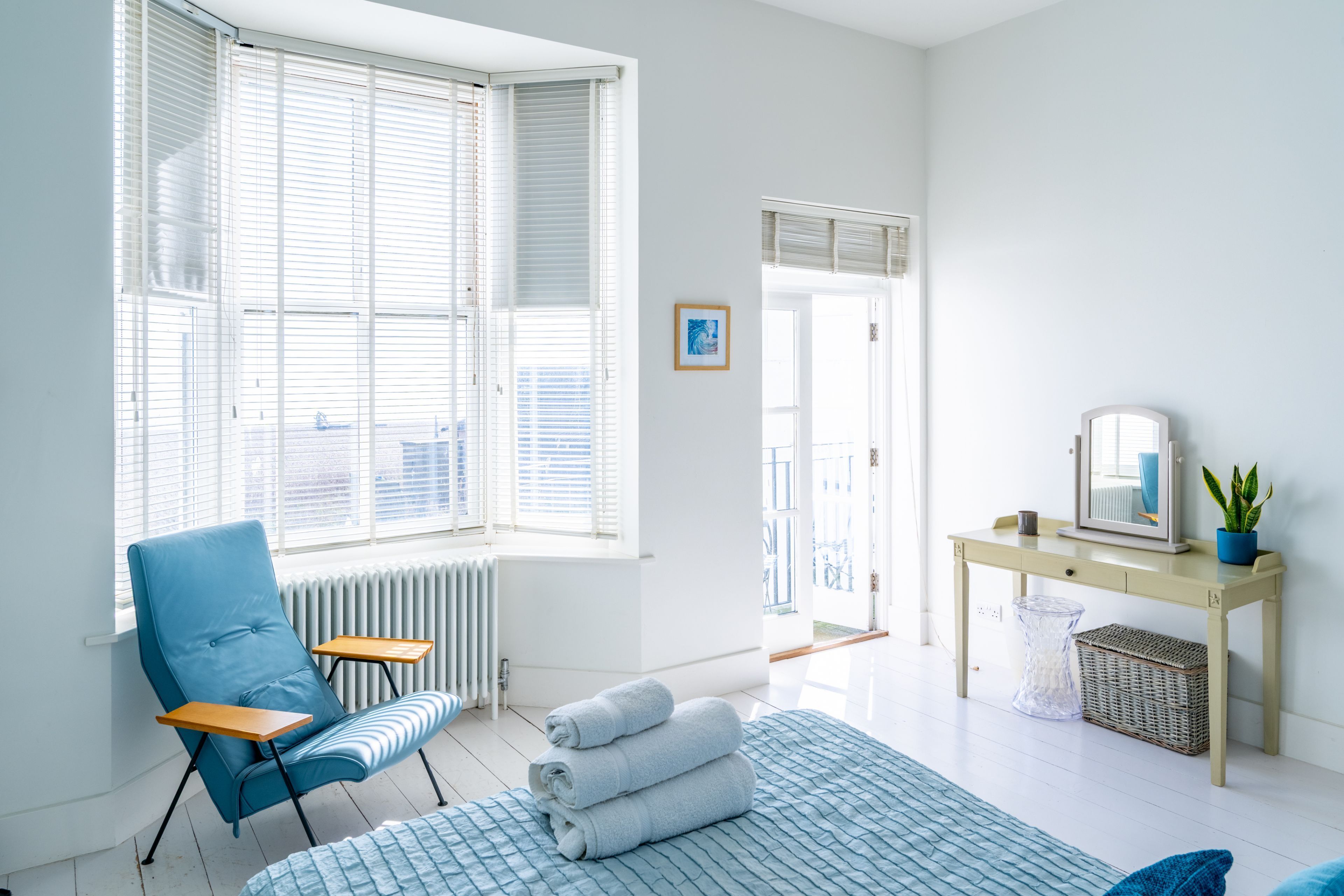 Quarto moderno com uma janela ampla, uma cadeira azul, uma cama com colcha azul, uma mesinha, um espelho e um vaso com planta.