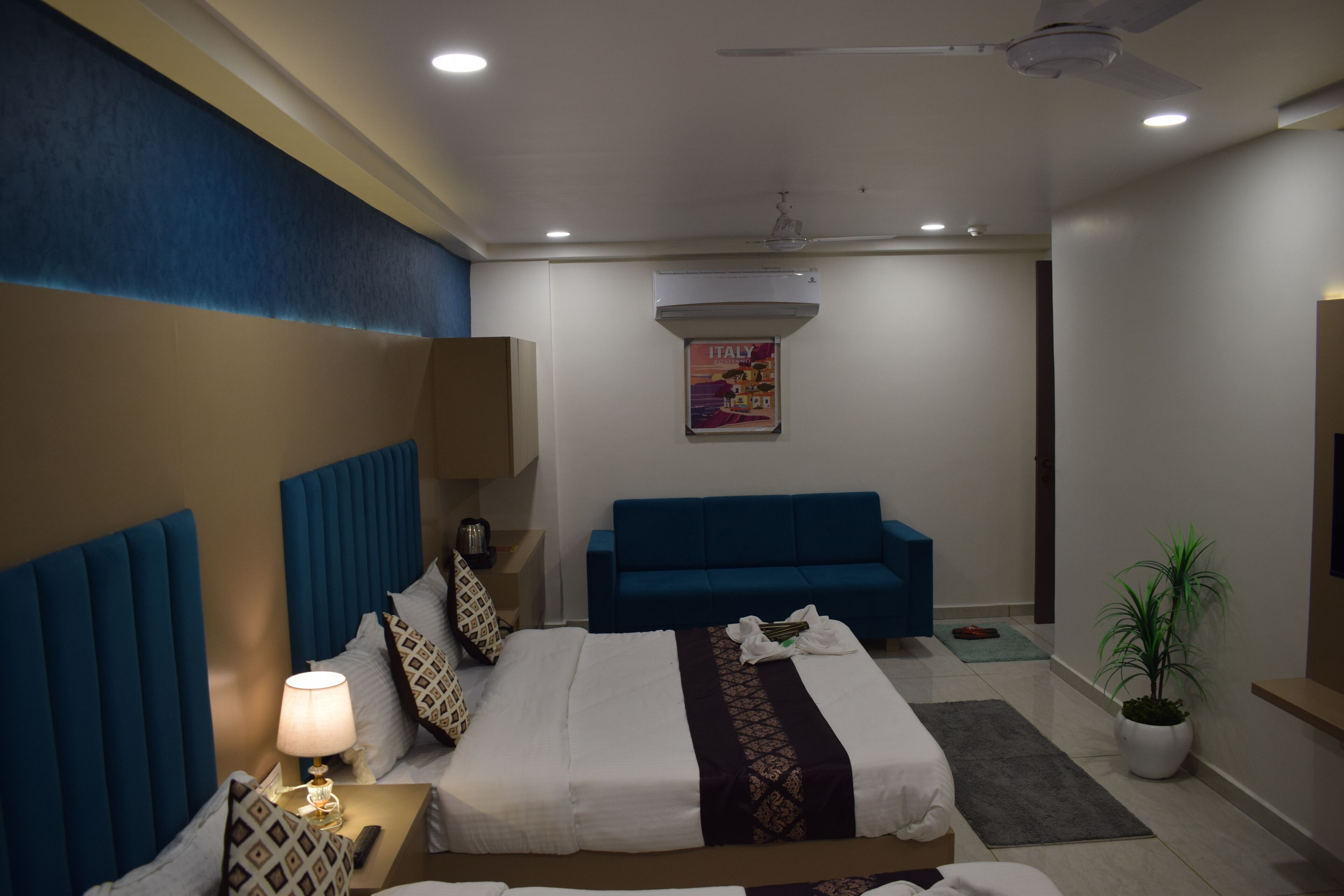A hotel room with a bed, a blue sofa, a ceiling fan, an air conditioner, and a potted plant.