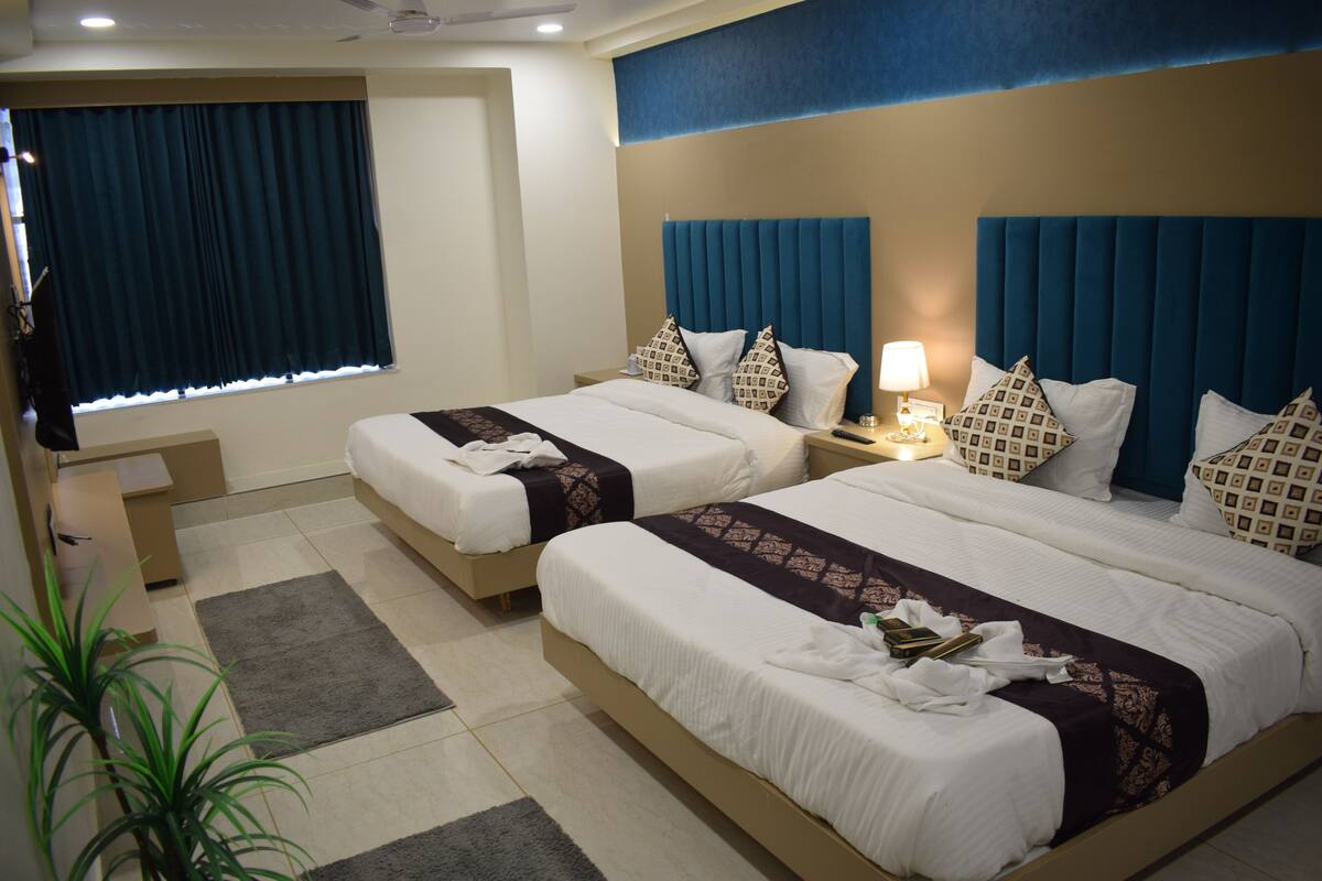 A hotel room with two beds, a desk, a chair, a window with curtains, and a potted plant.