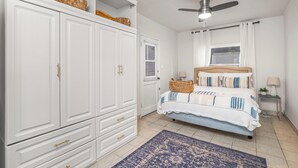 Een slaapkamer met een bed, een kledingkast, een plafondventilator, een raam met gordijnen en een klein tafeltje met een lamp.