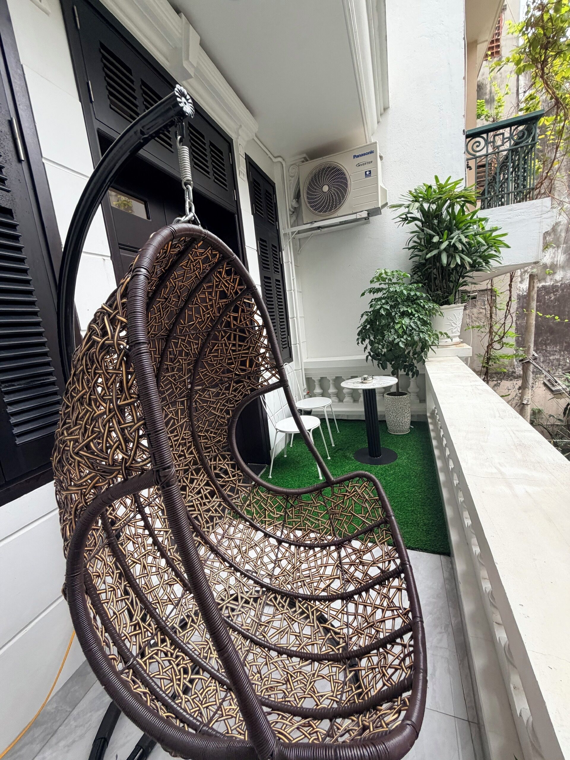Una silla colgante de mimbre con estructura metálica, una pared blanca con una ventana, una mesita con sillas y una planta en maceta.