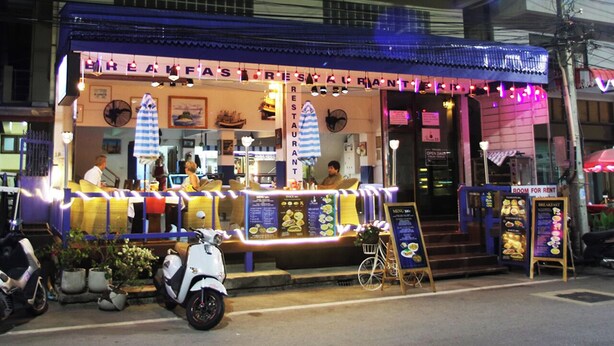 A brightly lit restaurant with outdoor seating, a menu board, and a sign indicating a room for rent.
