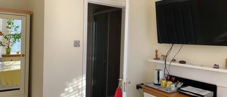 A compact hotel room with a shower, a bed, a TV mounted on the wall, and a small shelf with decorative items.