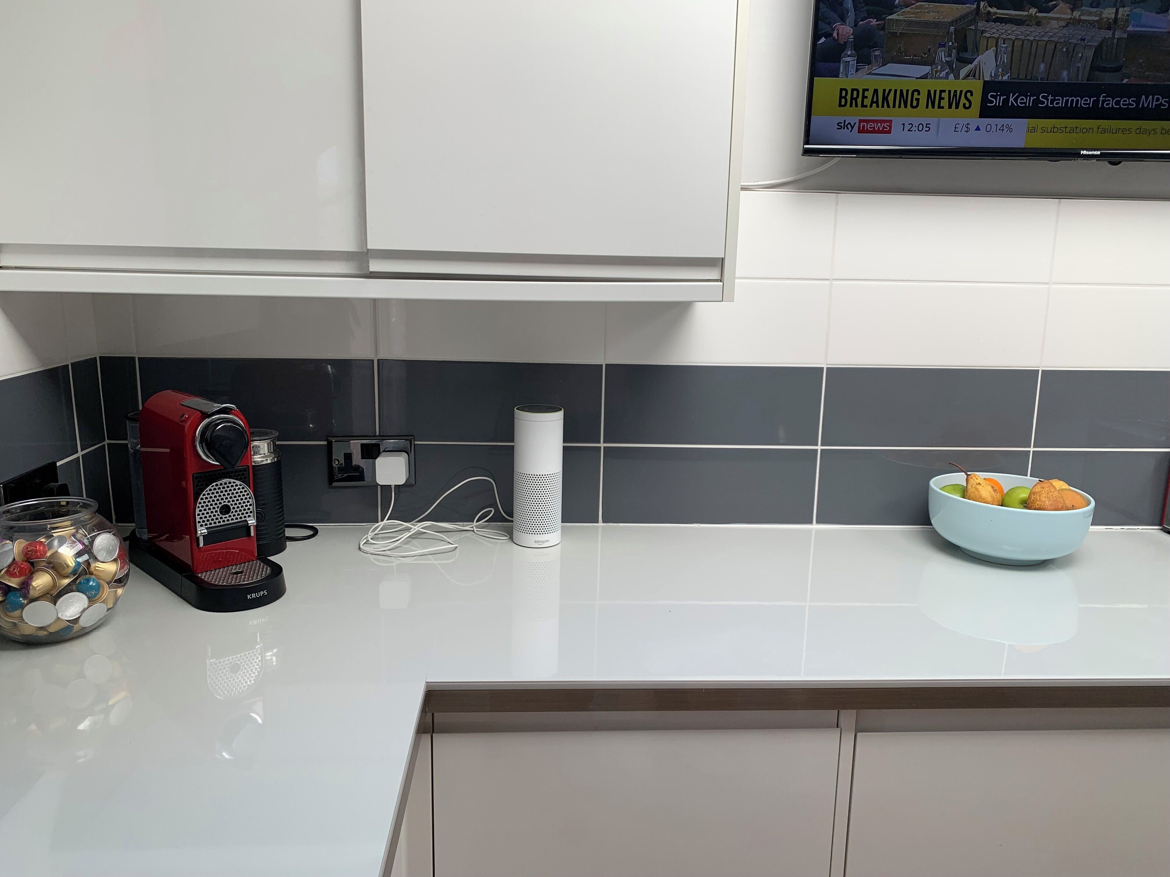 Et moderne køkken med kaffemaskine, en smart højttaler og en skål med frugt på bordet.