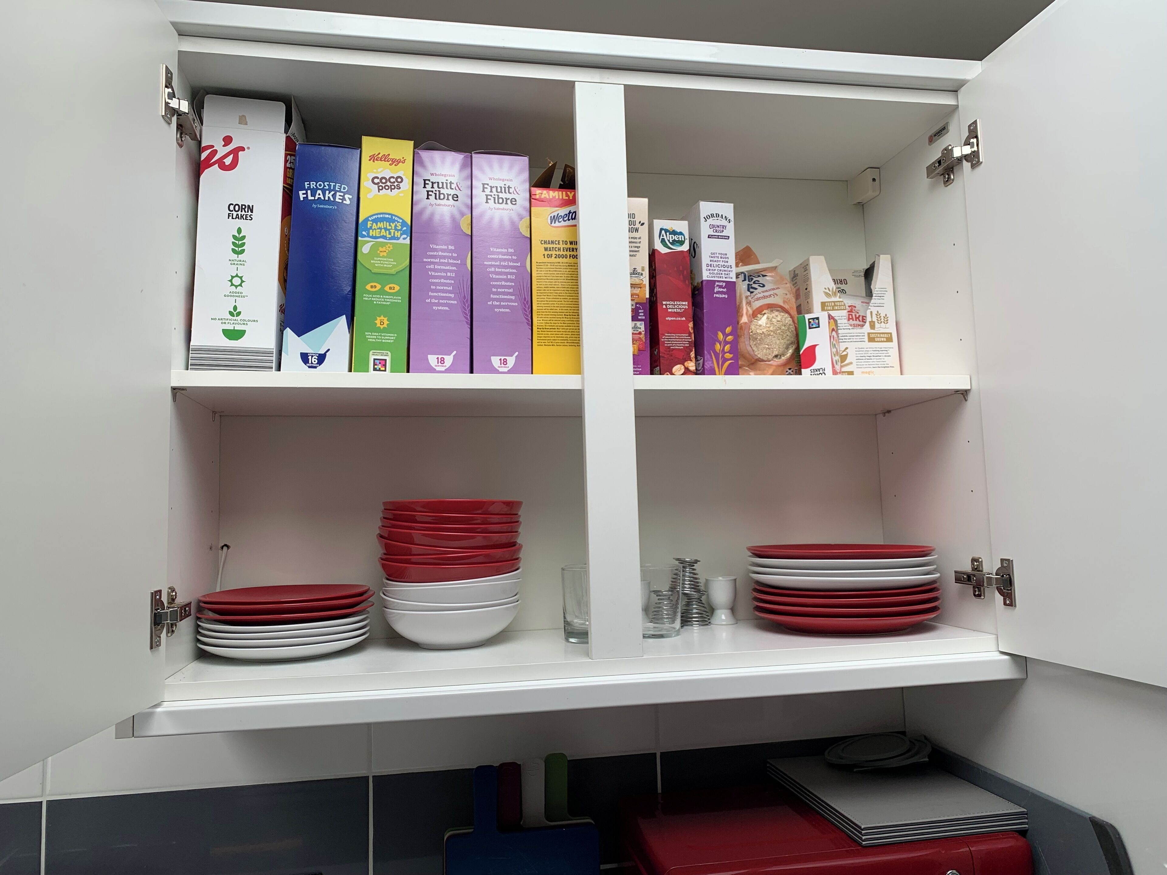 A kitchen cabinet with shelves holding cereal boxes, plates, and kitchen utensils.