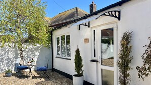 A white building with a sign reading "meads cabin," a small table with chairs, and potted plants. - Meads Cabin - Charmouth, Lyme Regis (Charmouth)